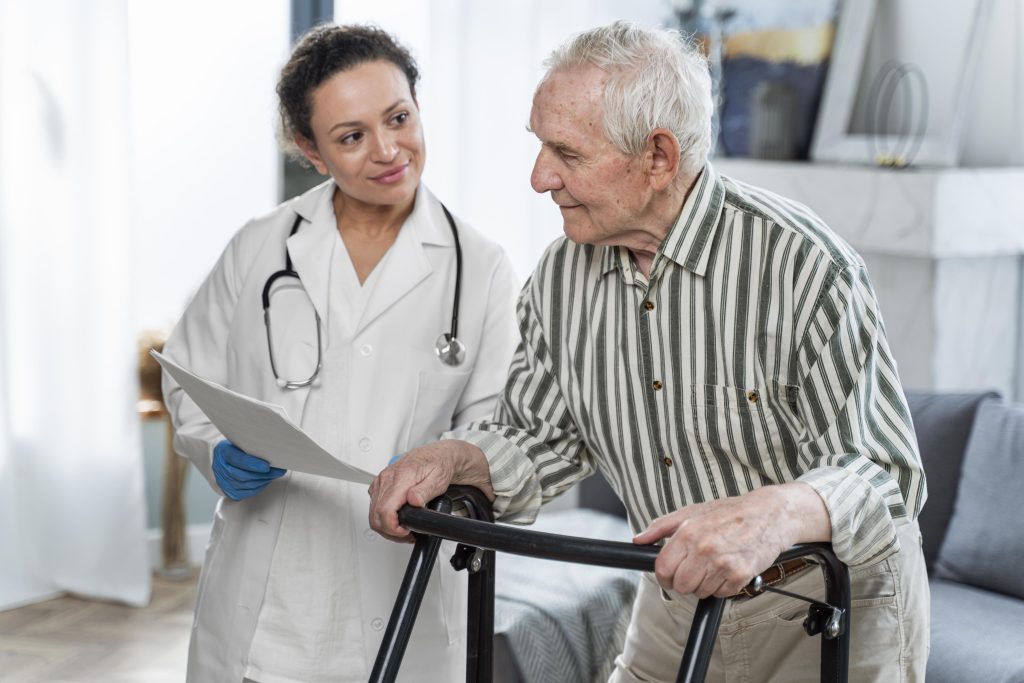 Profissional de saúde auxilia idoso próximo a uma cadeira de rodas, simbolizando o cuidado essencial em casos de Alzheimer e demência.