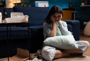 Mulher com expressão de tristeza sentada no chão da sala, abraçando um travesseiro, cercada por embalagens de comida e ambiente desorganizado.